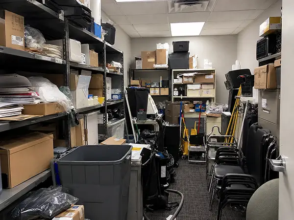Cluttered commercial office storage room before organization with crowded shelves, boxes, supplies, and blocked workspace